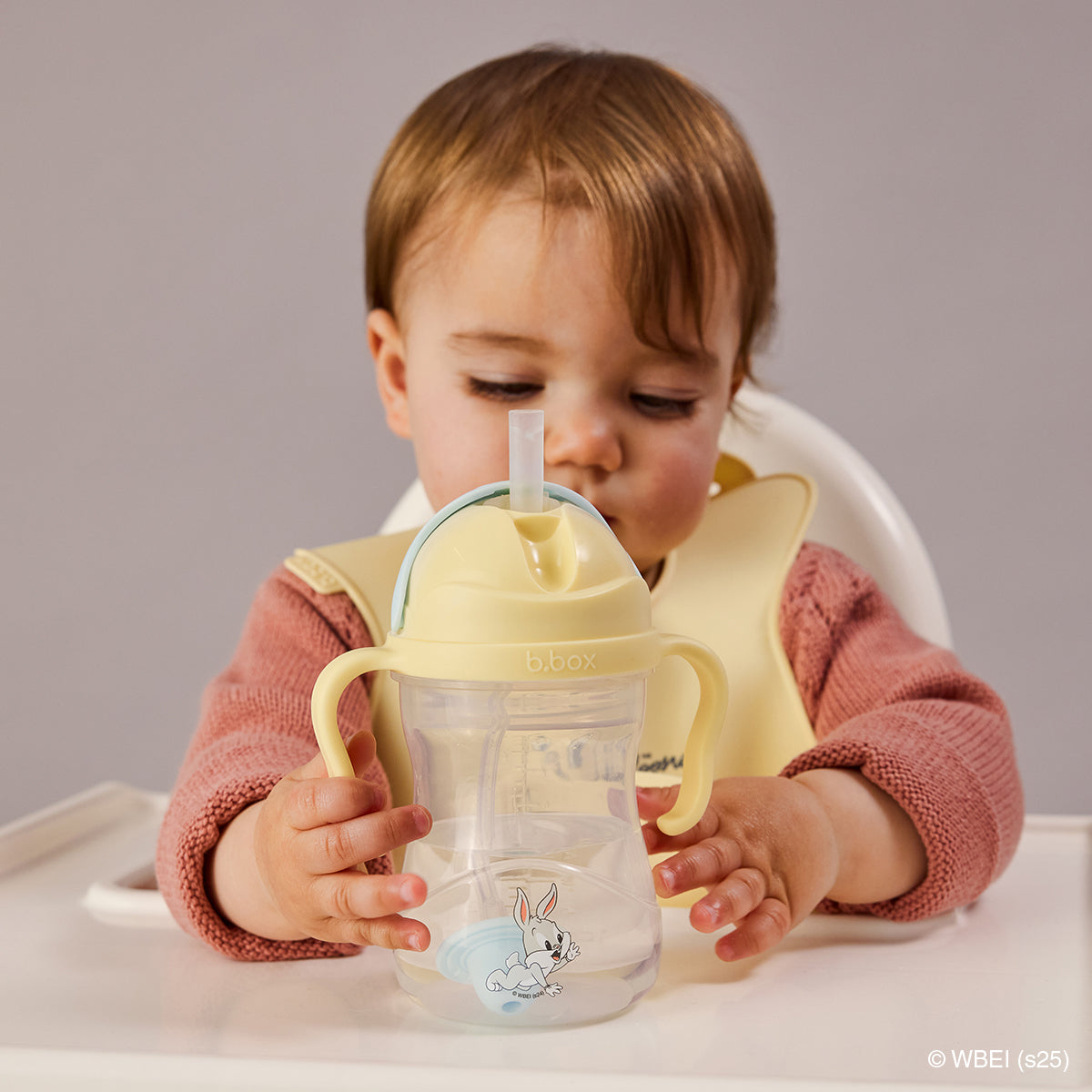 Child holding a yellow sippy cup with a Bugs Bunny design, sitting in a high chair against a plain background.