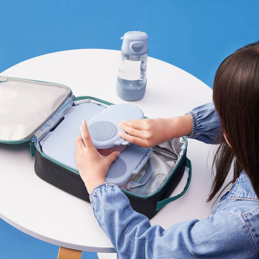A person in a denim jacket opens a black and teal lunch bag on a white table, placing a light blue insulated container inside. A clear water bottle with a blue lid sits nearby against a solid blue background.