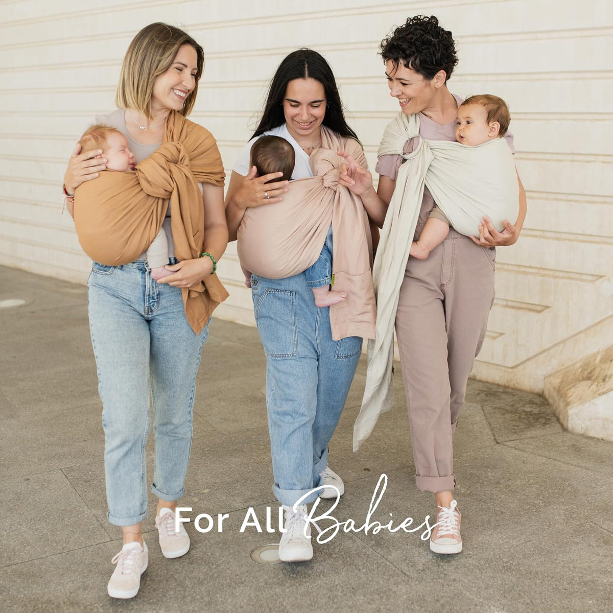 Three women walking outdoors while wearing baby wrap carriers in different colors: tan, blush pink, and cream. The image includes a text overlay at the bottom that reads 'For All Babies'.