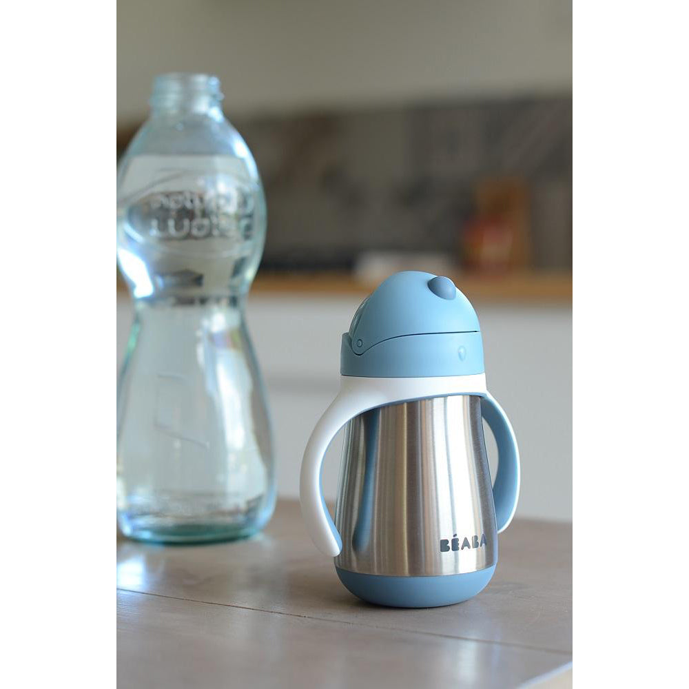 Blue and silver insulated sippy cup on a kitchen counter with a blurred water bottle in the background.
