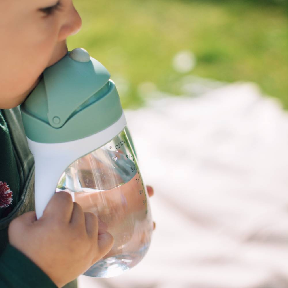 A close-up lifestyle shot of a child drinking from a clear plastic water bottle with a mint green and white flip-top lid, set against a soft-focus outdoor background.