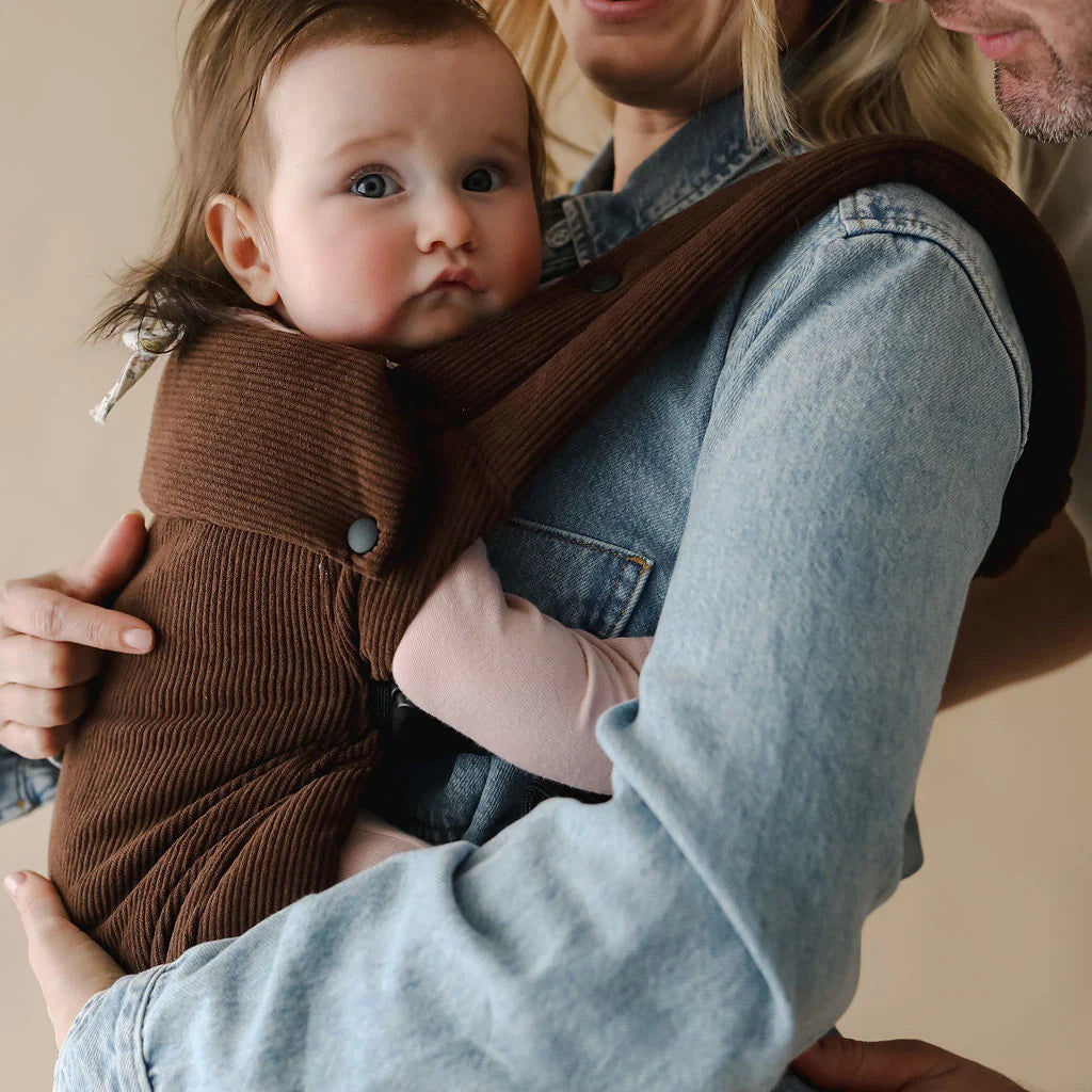 Person holding a baby in a brown fashion baby carrier Boba Auri against a neutral background