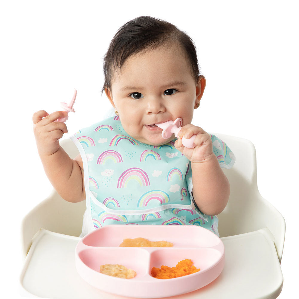 A baby with dark hair in a blue rainbow bib sits in a white highchair holding a chewtensil cutlery spoon and fork. A pink  bumkins grip dish is attached to the tray with food inside.