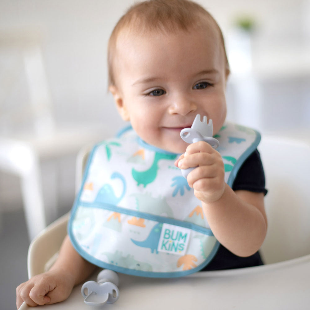 A child with brown hair in a blue dinosaur bib and black shirt sits in a white highchair holding a chewtensil cutlery fork to their mouth. A grey chewtensil spoon is laid out on the tray