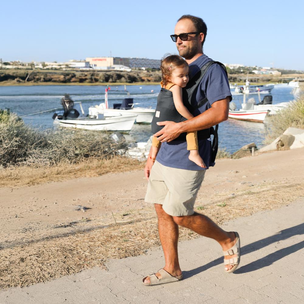 Man at the beach carrying baby on ttheir front in a mesh travel carrier