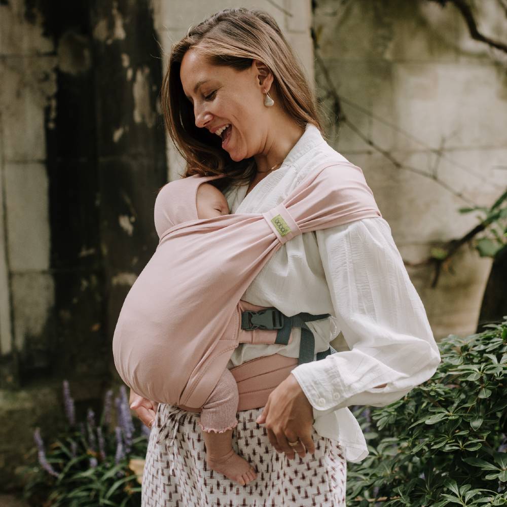 Woman holding a baby in a pink Bliss Hybrid Baby Carrier outdoors.