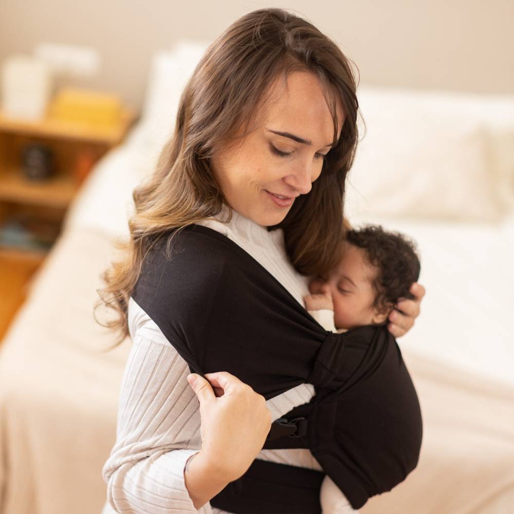 Woman holding a baby in a black Boba Bliss Hybid Carrier wrap sling on a bed.
