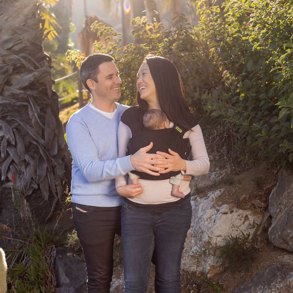 Couple with a baby in a carrier standing in a natural setting with greenery and rocks.