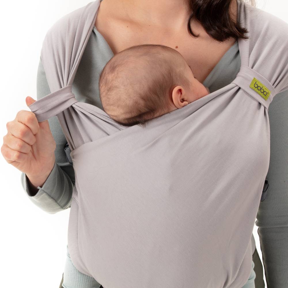 Person holding a baby in a gray baby carrier with a visible brand label.