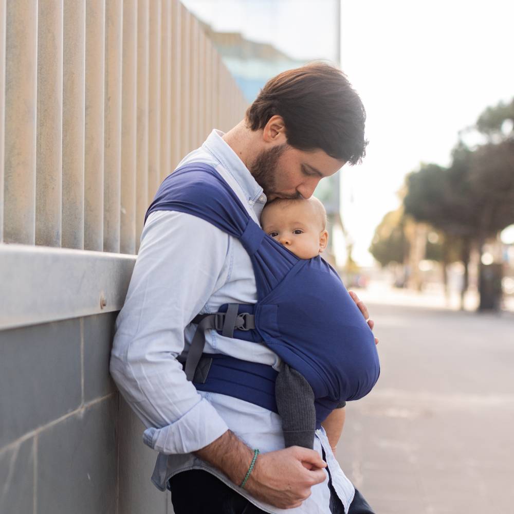 Man holding a baby in a blue carrier on a city street.