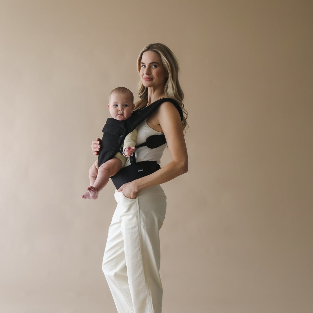 on trend baby carrier worn forward facing by a woman holding a baby in a carrier against a beige background