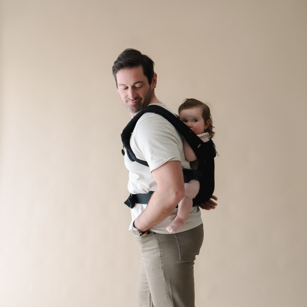 Man holding a baby in a carrier against a plain background, baby in worn on the back