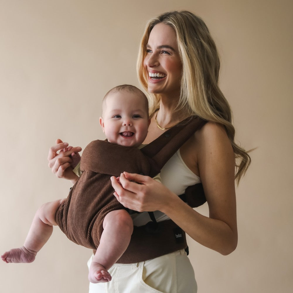 Woman holding a baby in a brown carrier in the front facing position against a beige background