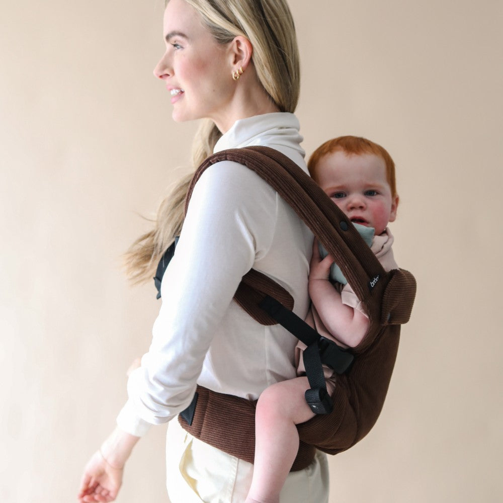 Person wearing a baby in a brown carrier in the back carrying position against a beige background