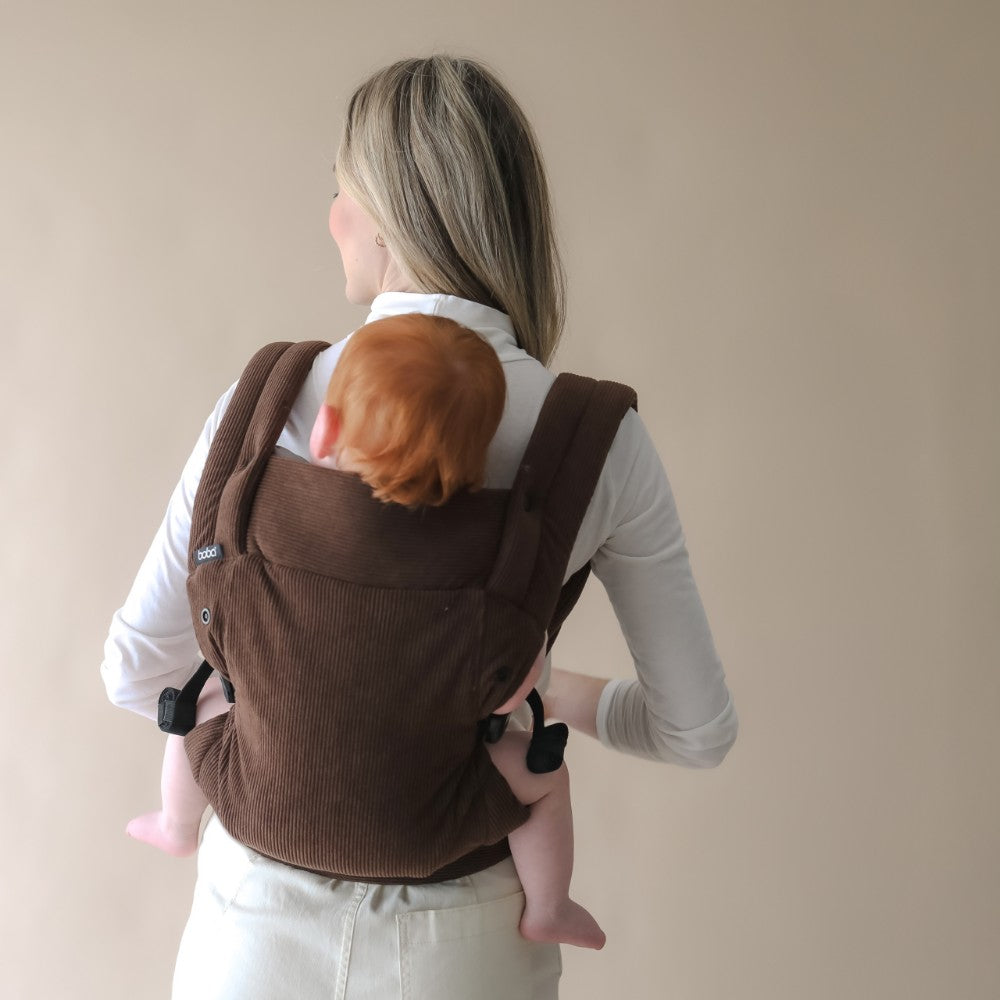 Person wearing a baby in a brown carrier in the back carrying position against a beige background