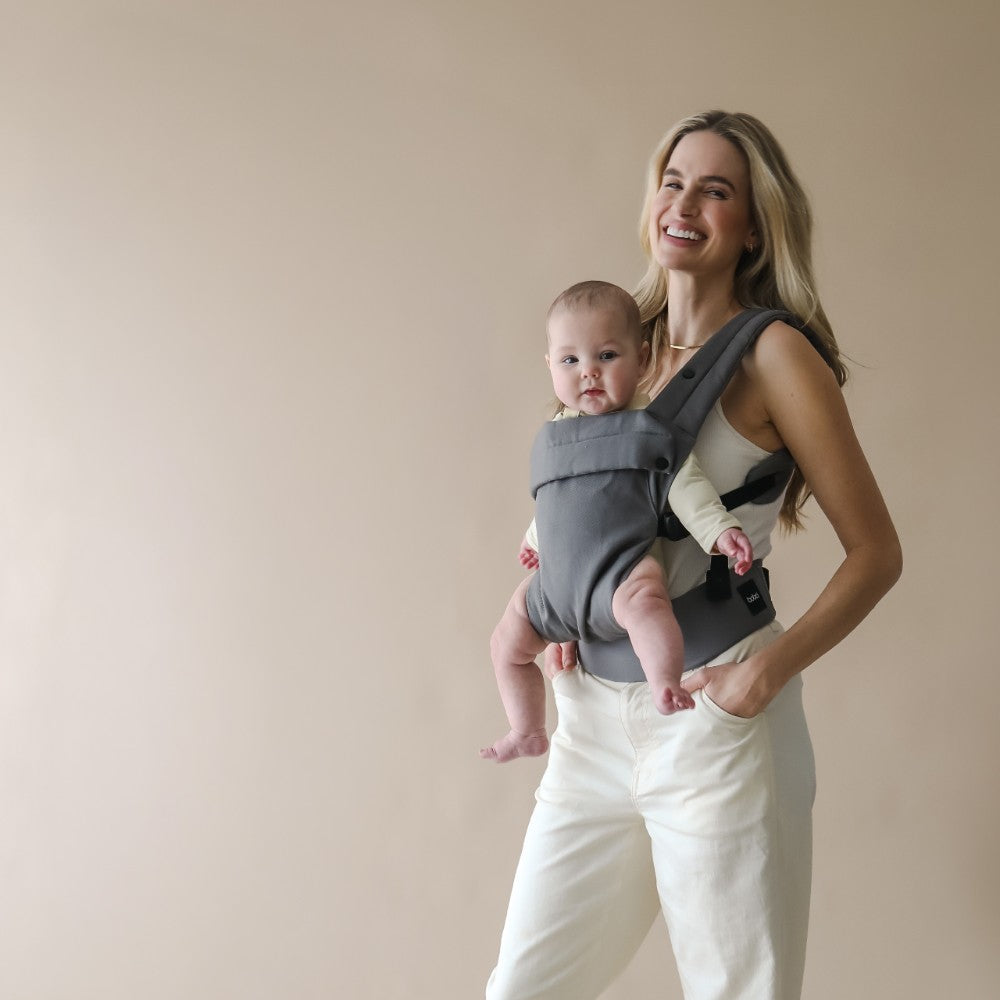 Woman holding a baby in a gray Boba Auri carrier in the front facing position against a beige background