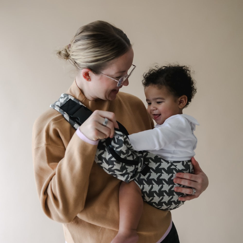 Woman holding a child in a patterned baby carrier against a plain background