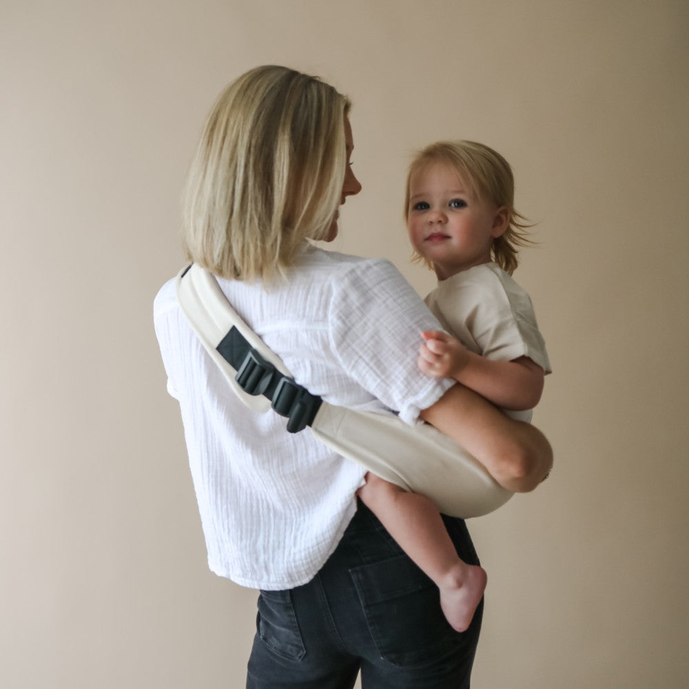 Woman carrying a child in a toddler carrier worn on the hip against a plain background