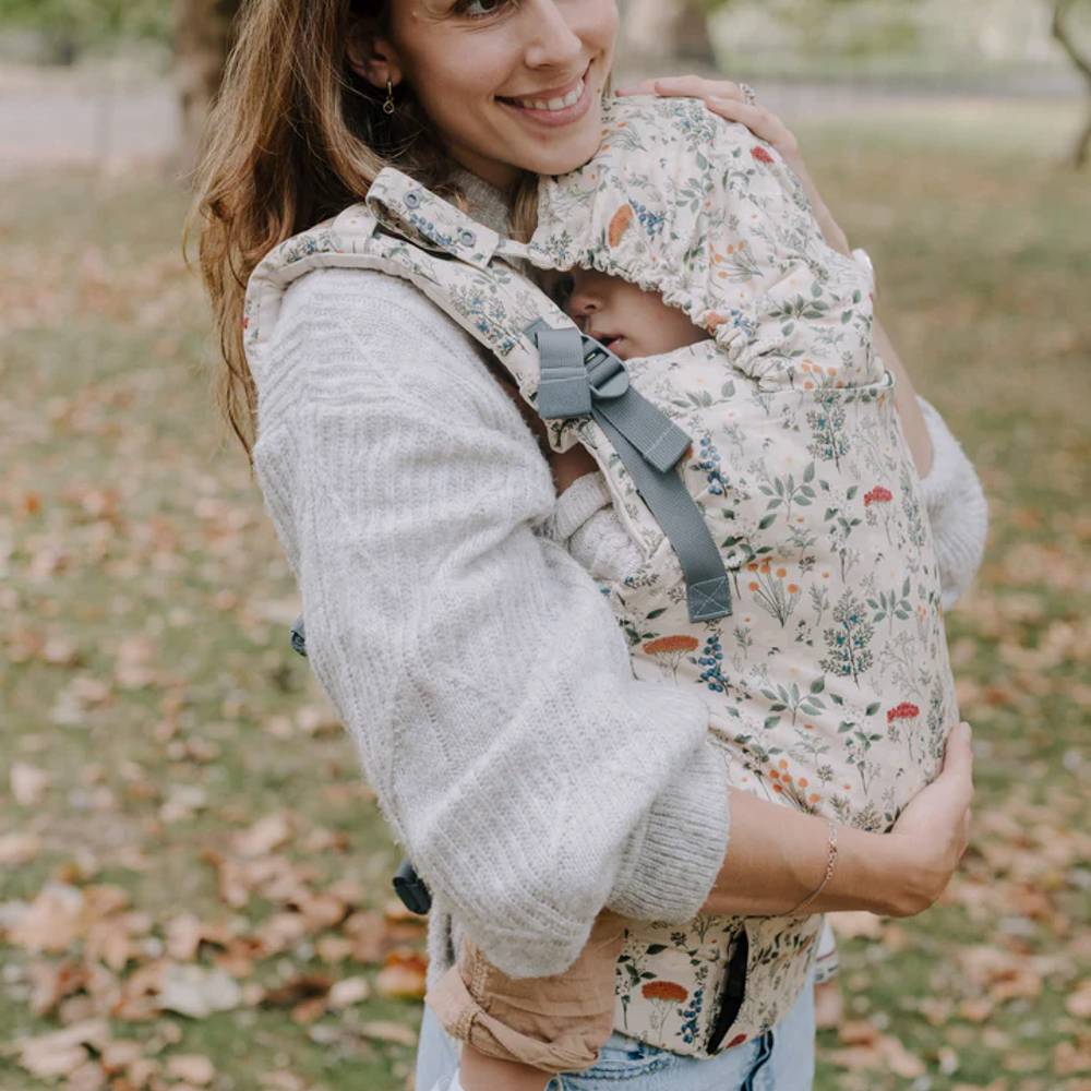 Woman carrying a child in a floral baby carrier outdoors on a grassy area with fallen leaves.