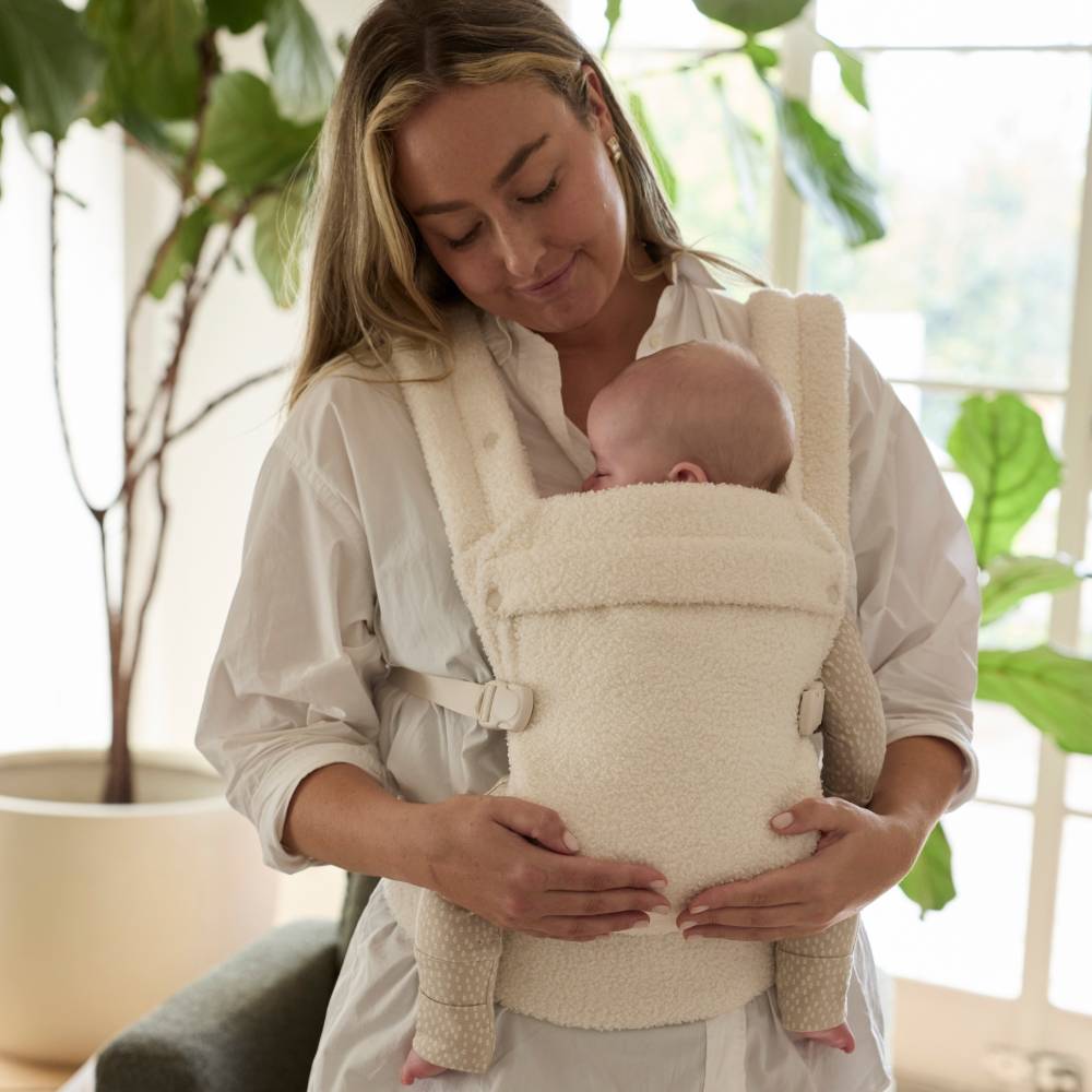 Woman holding a baby in a beige boucle baby carrier with plants in the background