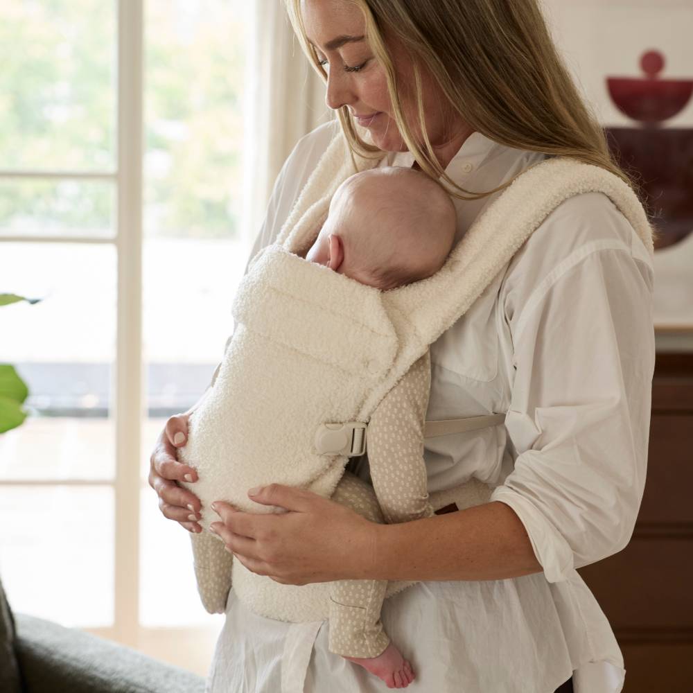Woman holding a baby in a beige baby carrier indoors.