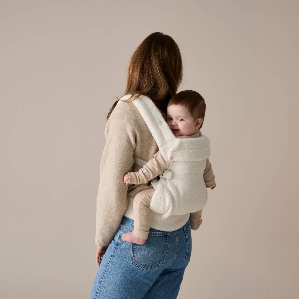 Woman carrying a baby in a white baby carrier against a beige background