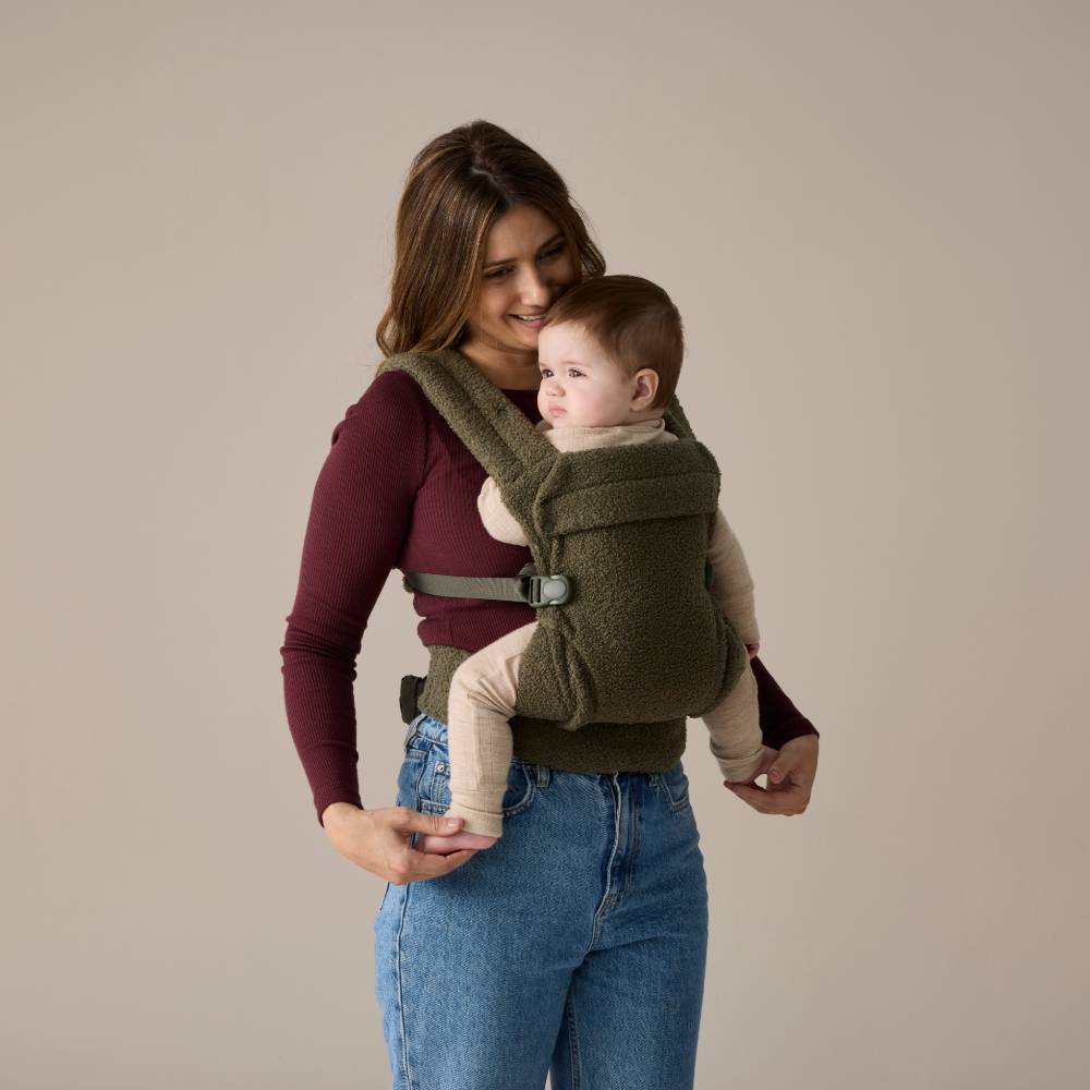 Woman holding a baby in a green baby carrier against a beige background