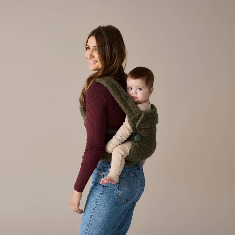 Woman carrying a baby on her back in a green Boba Auri Teddy baby carrier against a beige background