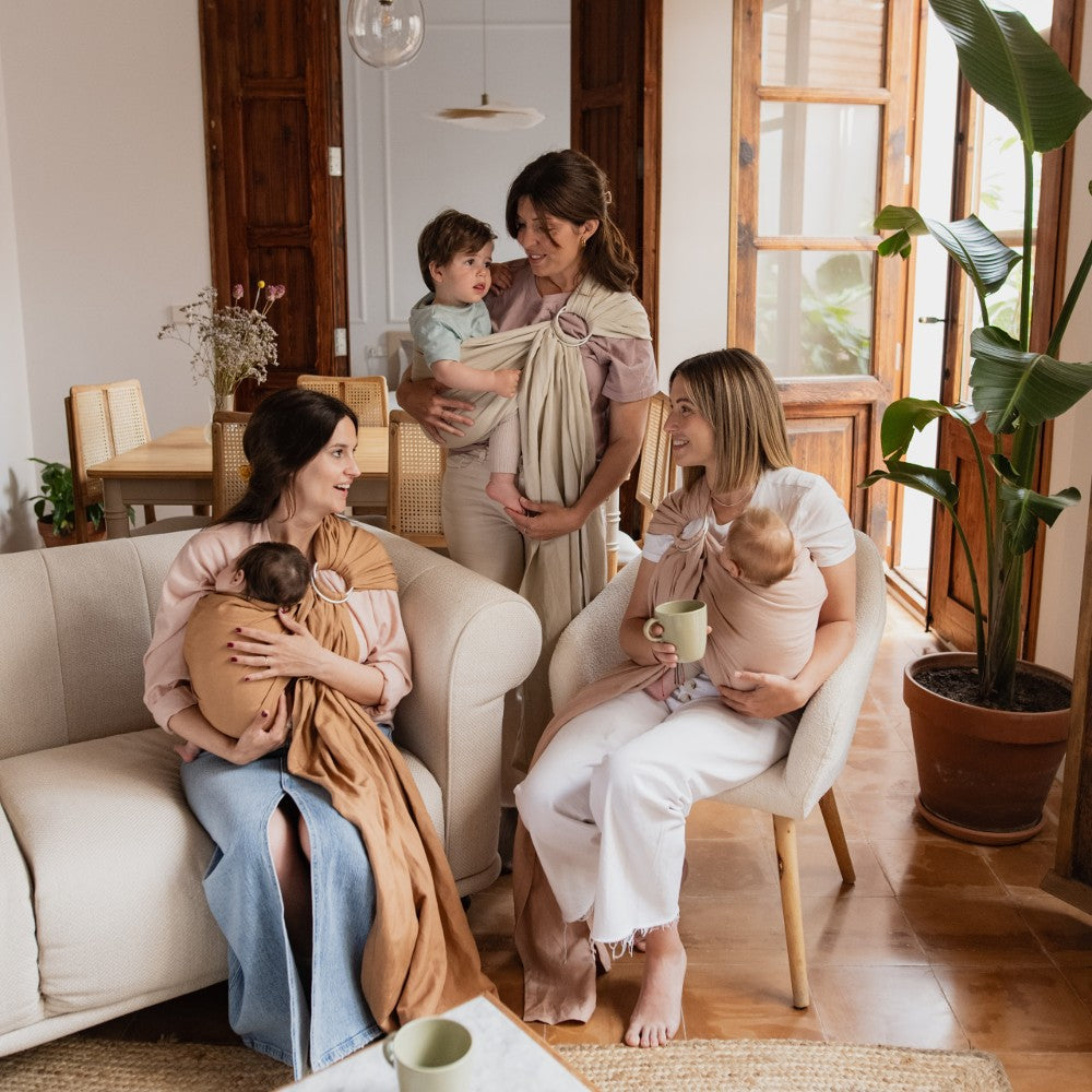 Three women sitting in a living room with their babies using wraps, holding cups.