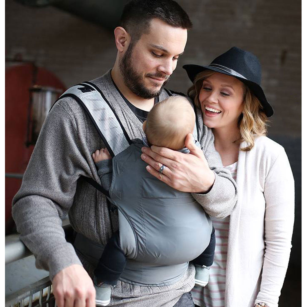 Man holding a baby in a travel carrier with a woman standing next to them, all smiling.