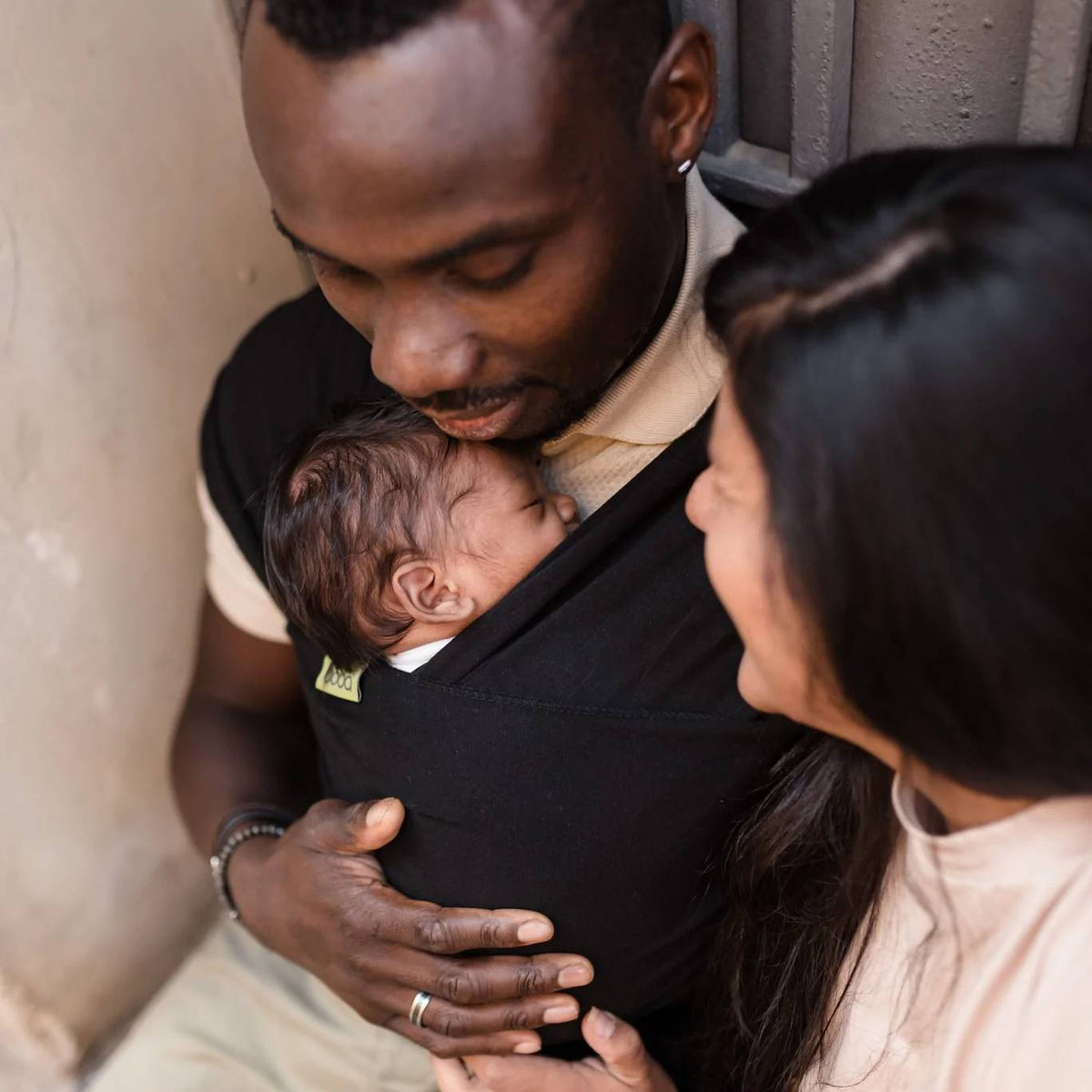 Man holding a baby in a sling with a woman standing next to them, all indoors.