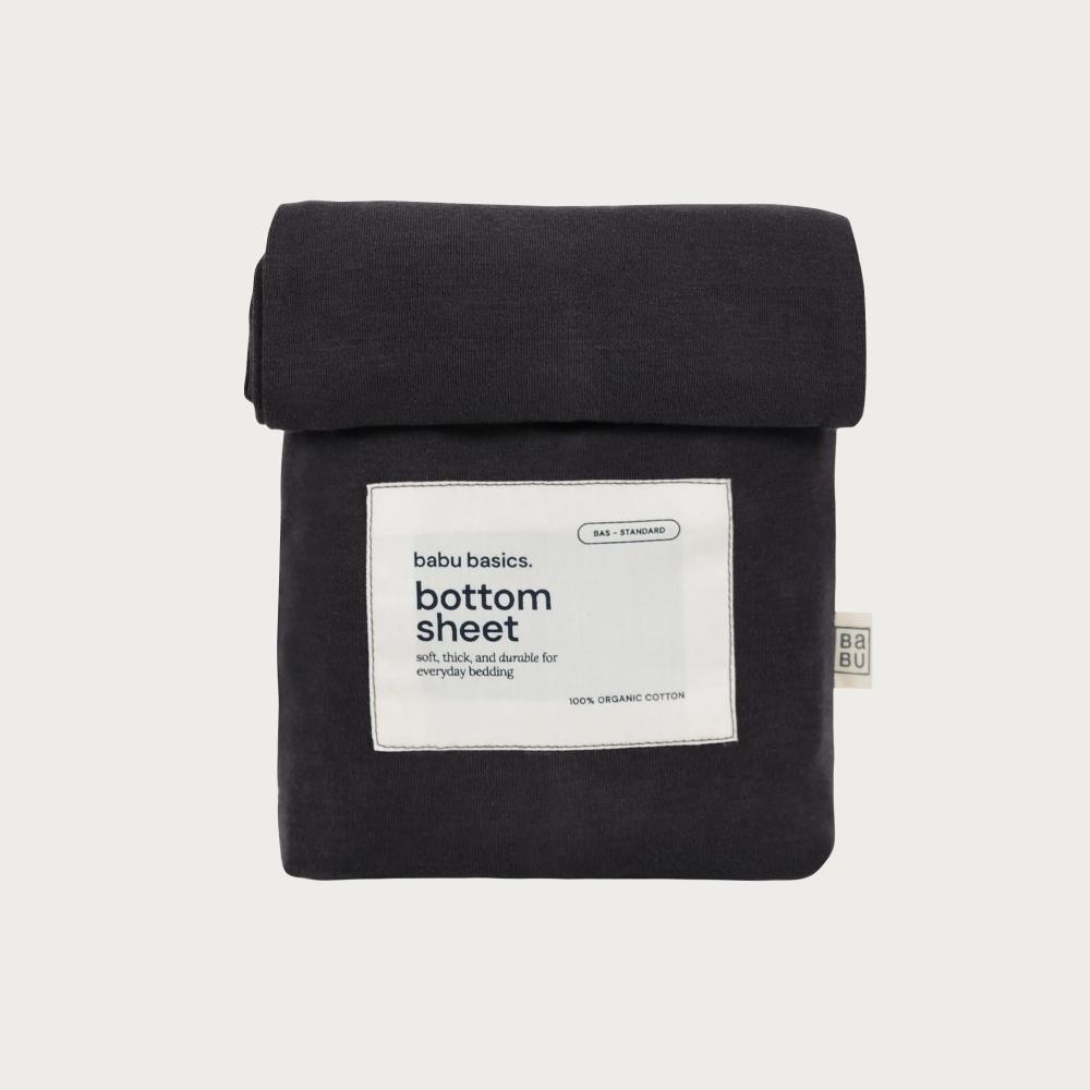 A folded black cotton bottom sheet with a white paper label reading 'babu basics bottom sheet' on a light gray background.