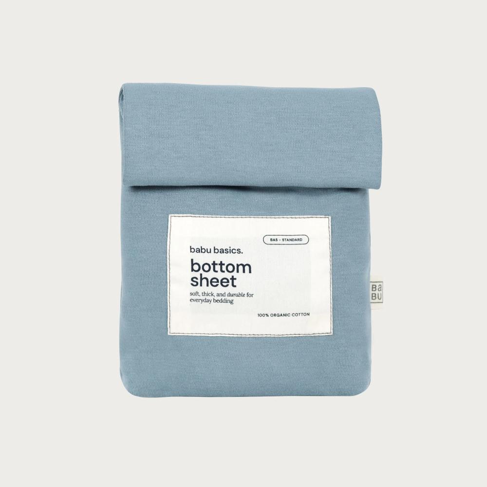 A folded light blue cotton bottom sheet with a white paper label reading 'babu basics. bottom sheet' on a light gray background.