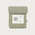 A sage green cotton bottom sheet by babu basics, neatly folded and packaged in a matching fabric sleeve with a white paper label on the front, set against a solid light gray background.