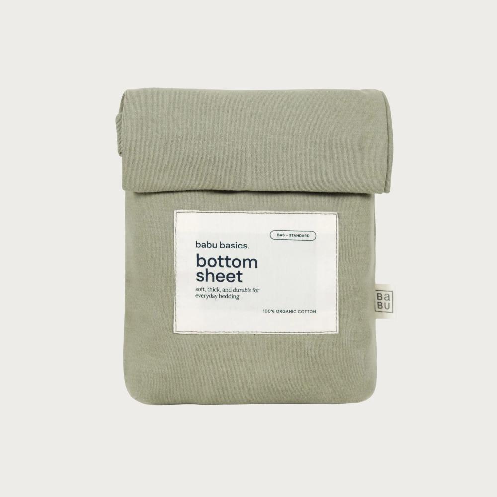 A sage green cotton bottom sheet by babu basics, neatly folded and packaged in a matching fabric sleeve with a white paper label on the front, set against a solid light gray background.