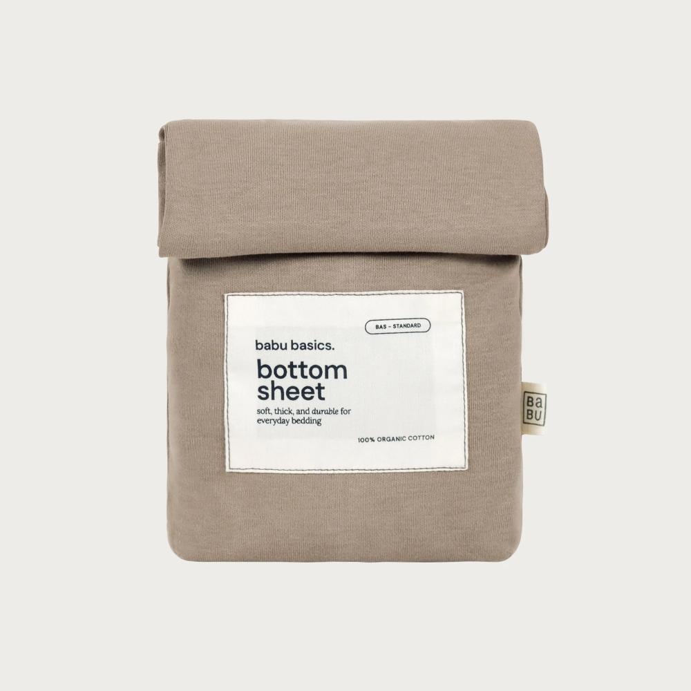 A folded beige cotton bottom sheet with a white paper label reading 'bubu basics. bottom sheet' and a small side tag, photographed on a light gray background.