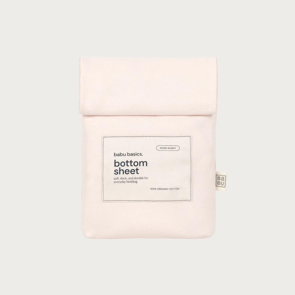 A pale blush pink cotton bottom sheet by babu basics, neatly folded and packaged in a matching fabric sleeve with a white paper label, set against a solid light gray background.