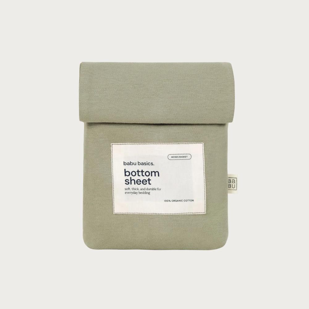 A sage green bottom sheet by babu basics, neatly folded and packaged in a matching fabric sleeve with a white paper label, set against a solid light gray background.