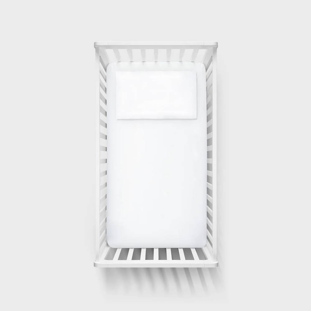 A top-down view of a white wooden crib with a white mattress and pillow, set against a solid light gray background.