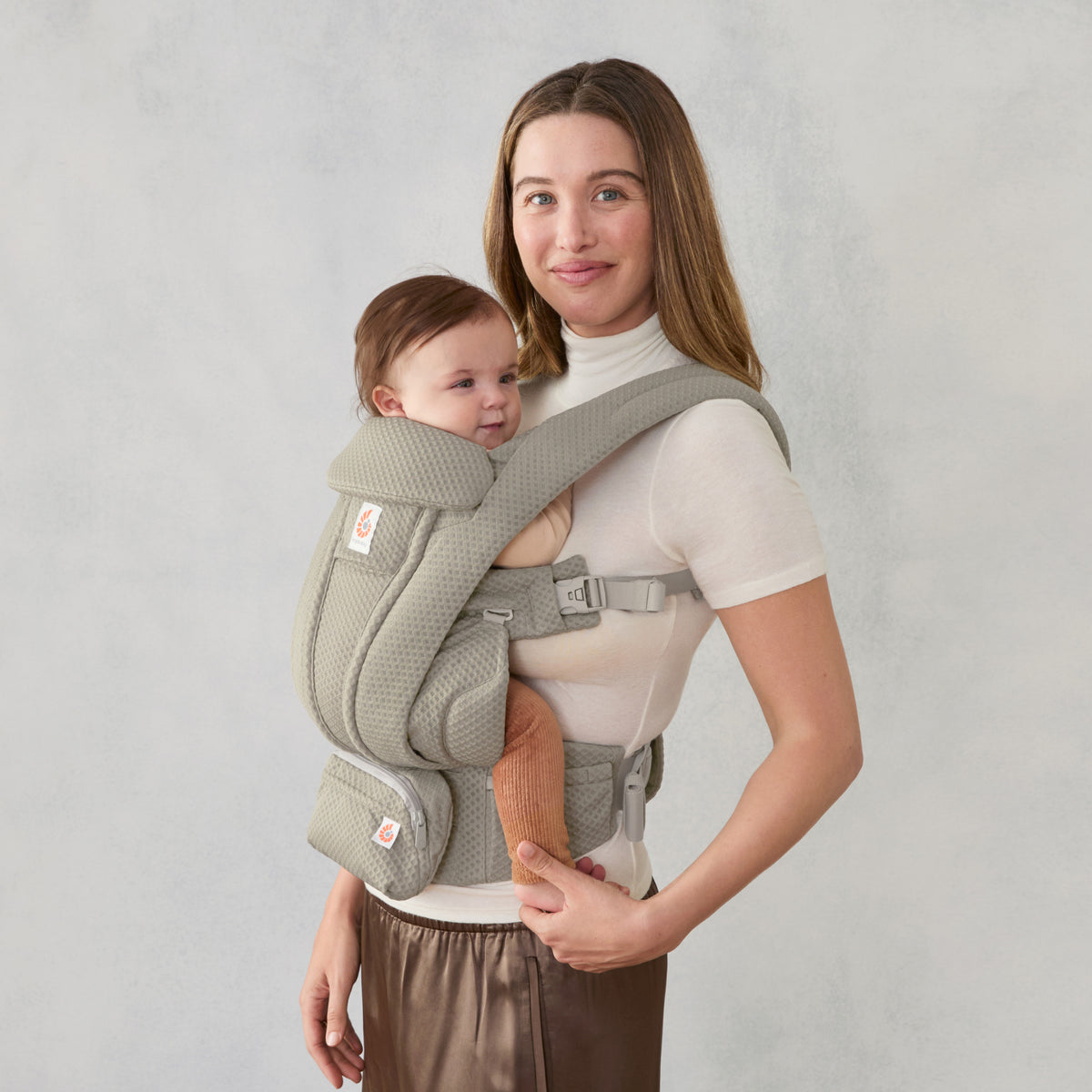 A side view of a woman wearing an olive green baby carrier with a baby inside on a light grey background. 