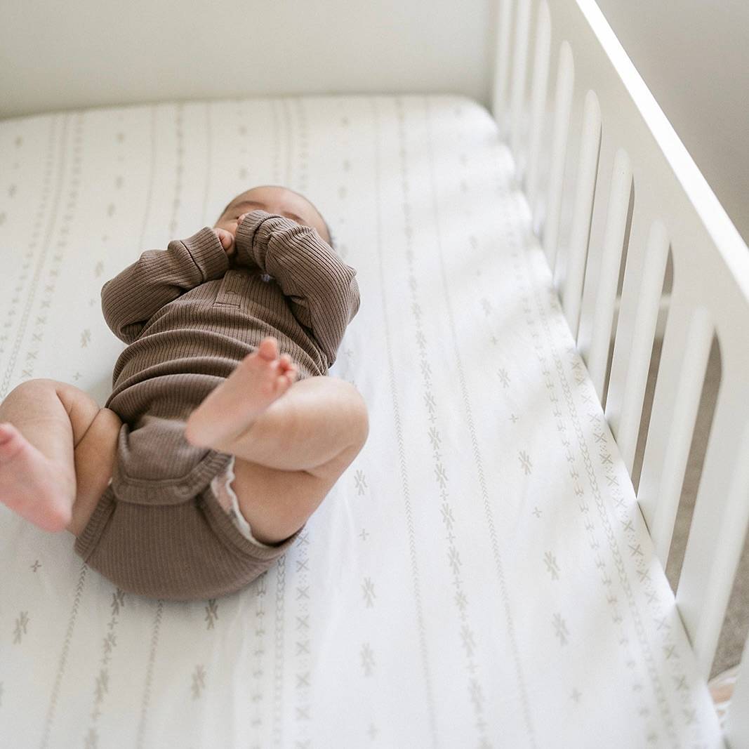 A baby in a cot on top of a pacific island inspired patterned cot sheet. 