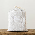A packaged fitted cot sheet. It is white with a grey maori inspired pattern. 