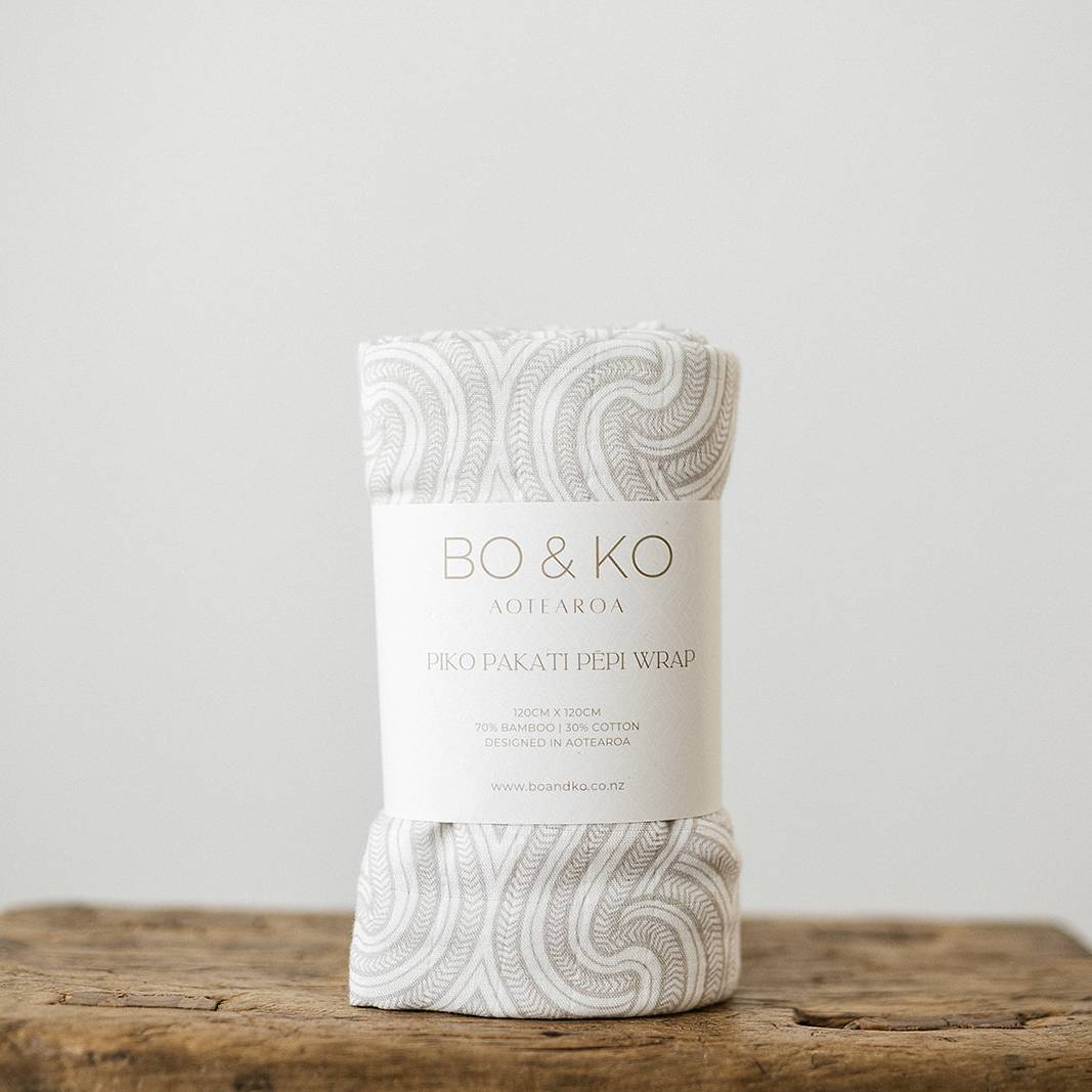 A white muslin baby swaddle with a grey maori inspired pattern on it. 