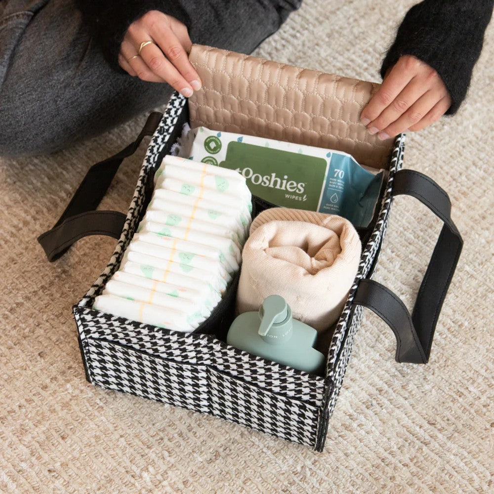 A black-and-white houndstooth baby diaper storage basket with black handles is shown open on a beige carpet, containing diapers, wipes, a folded blanket, and a bottle. A person's hands and legs are visible holding the basket open.