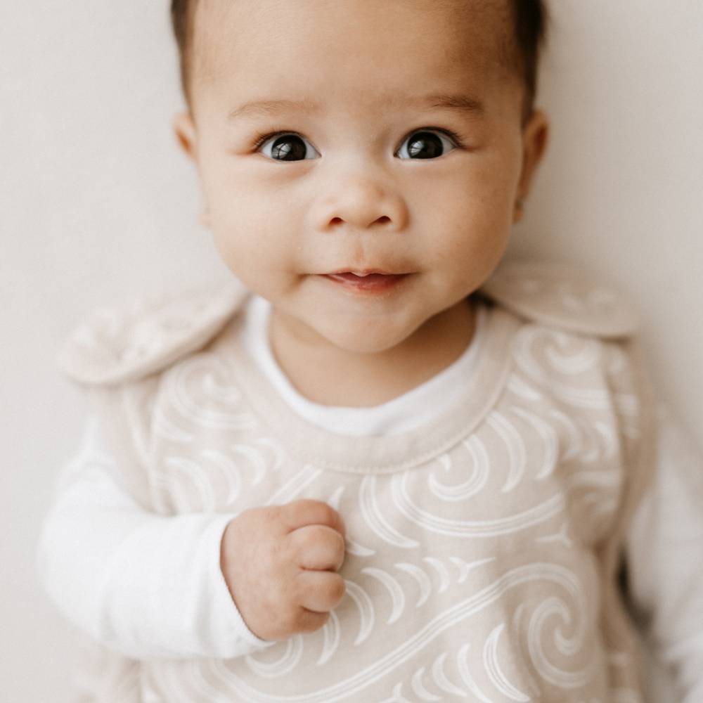 A close-up of a baby wearing a cream-colored long-sleeve top with a white maori pattern, photographed against a soft, light beige background.