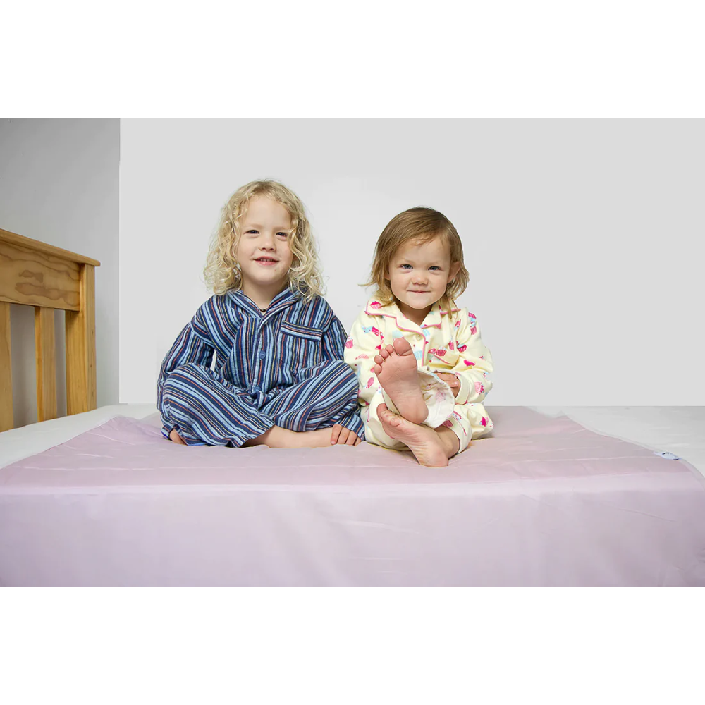 Two young children sitting on a bed wearing matching pajamas. The child on the left wears blue and white striped pajamas, and the child on the right wears a cream pajama set with a pink and yellow pattern. They are sitting on a dusty pink brolly sheet.