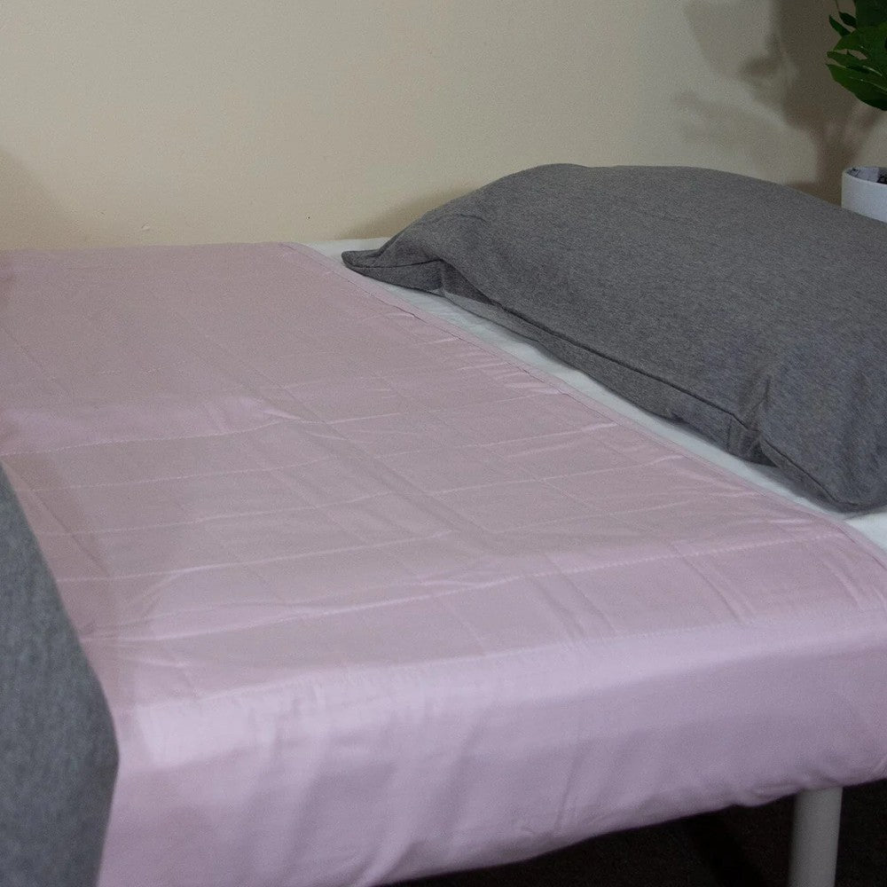 A light pink quilted brolly sheet on a bed with a gray pillow and matching gray blanket, shown in a bedroom setting.