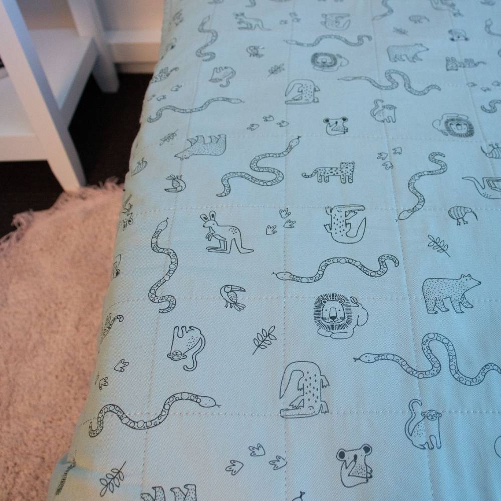 A close-up of a quilted brolly sheetin pale green with a repeating pattern of simple animal and snake illustrations in black, shown on a bed with part of a white chair and a pink rug visible.