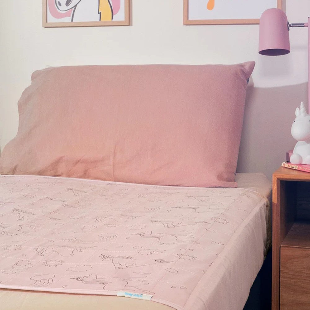 A pink bedding set featuring a solid pink pillowcase and a light pink quilted brolly sheet with delicate line-drawn unicorn patterns, styled in a bedroom setting next to a wooden nightstand and a pink lamp.