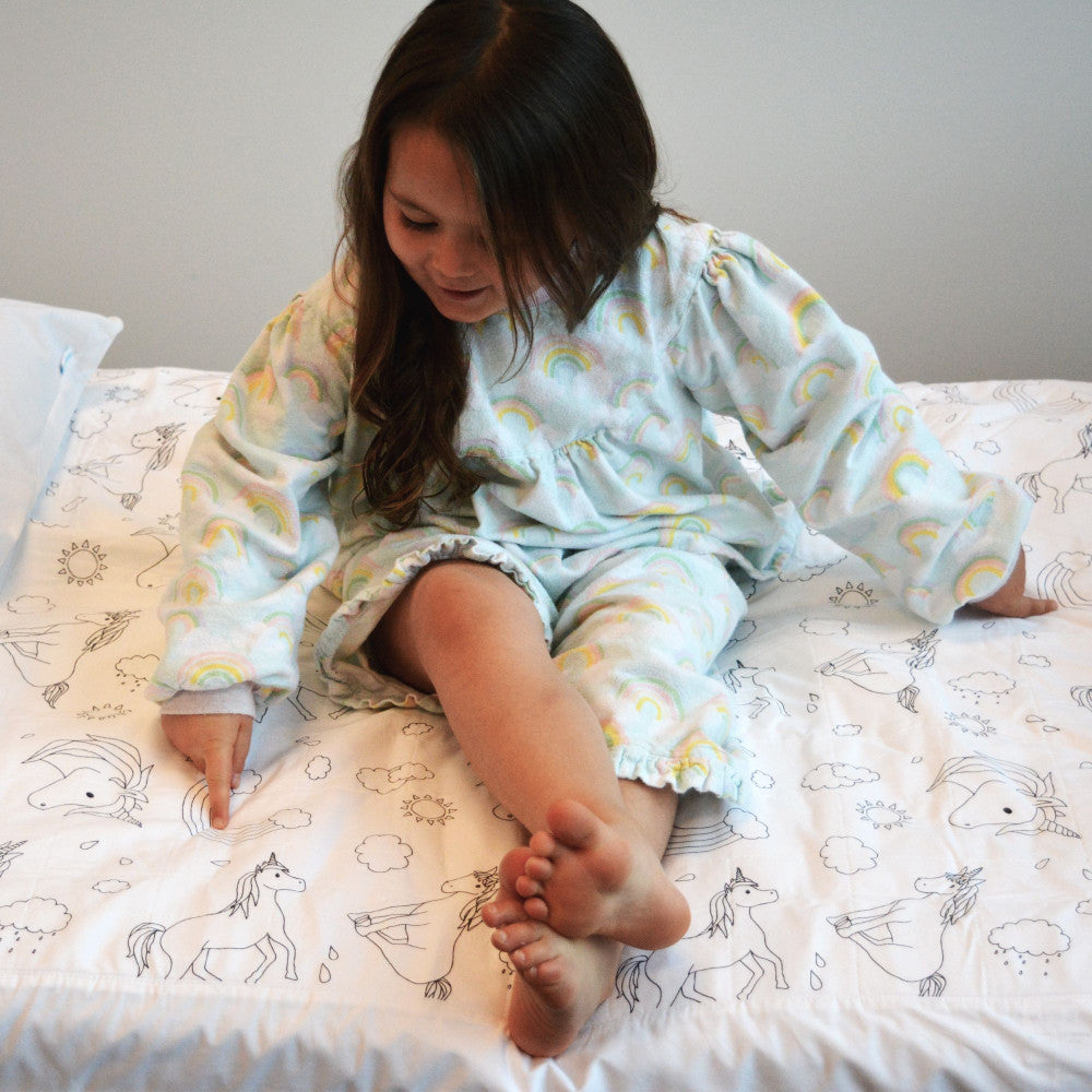 A young girl with long dark hair sits on a bed wearing a matching light blue pajama set with a rainbow and unicorn pattern. The bed is covered in a white sheet with black line drawings of unicorns and clouds.